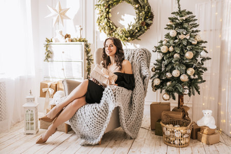 Beautiful cheerful happy young girl with christmas gifts on sofa on the background of a new year tree at homeの写真素材