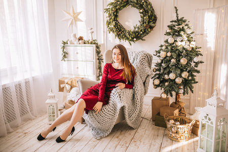 Beautiful cheerful happy young girl with christmas gifts on sofa on the background of a new year tree at homeの写真素材