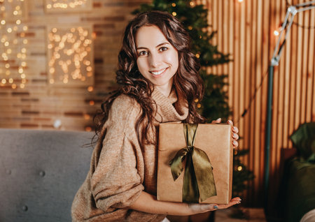 Beautiful cheerful happy young girl with christmas gifts on sofa on the background of a new year tree at homeの写真素材