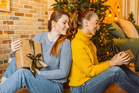 Two beautiful cheerful happy young girls girlfriend give Christmas gifts on the background of a new year tree at homeの写真素材
