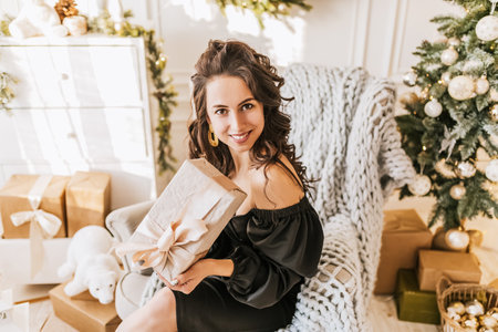 Beautiful cheerful happy young girl with christmas gifts on sofa on the background of a new year tree at homeの写真素材