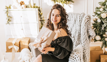 Beautiful cheerful happy young girl with christmas gifts on sofa on the background of a new year tree at homeの写真素材