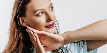A beautiful young girl smiles, dreams and holds her hands near her face on a white isolated gray background. Place for advertising. Positive woman with fashion make-up with purple lips.の写真素材