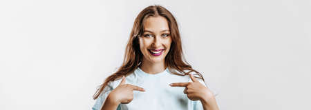 A beautiful young girl smiles and points her fingers at herself on a white isolated gray background. Place for advertising. Positive woman with fashion make-up with purple lips.の写真素材