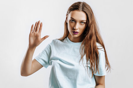 Hey. Beautiful young girl with a gesture greets, raises her hand with her palm to the camera on an isolated gray background. Brunette woman with fashion makeup with purple lips seriously lookingの写真素材