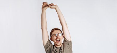 Good morning. The sleepy man yawns, pulls his arms up and stretches. Boredom and sleepiness. Young handsome man with glasses a brunette beard posing on gray background. Place for advertising.の写真素材