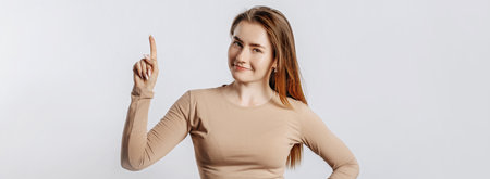 Beautiful young girl smiling and pointing her finger to the side at on a white isolated background. A woman points to an idea, a place for advertising. Positive brunette in a beige jumper.の写真素材