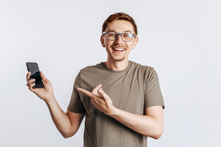 Young handsome man holding a smartphone and smiling. The guy cheerfully points his finger at the phone. Student, worker posing for photo isolated on gray background. Place for advertising.の写真素材