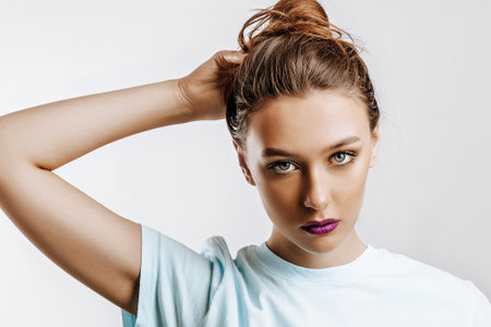 A beautiful brunette girl holds her hair and makes her own hairstyle. Young model with long hair, fashion makeup with purple lips on an gray background. The woman seriously looking at the camera.の写真素材