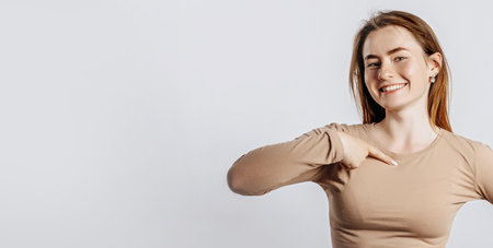 Beautiful young girl smiling and pointing her her finger at herself on a white isolated background. A woman points to an idea, a place for advertising. Positive brunette in a beige jumper.の写真素材