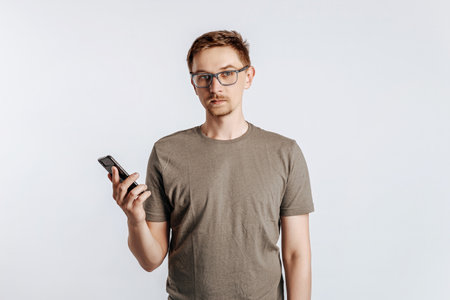 Young handsome man with glasses holding smartphone. Student, worker looks data information, posing for photo isolated on gray background. Place for advertising.の写真素材