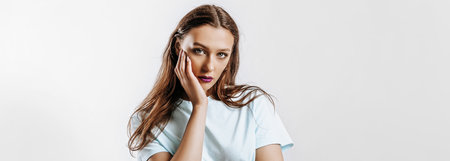 Beautiful young brunette girl with green eyes with long hair with fashion makeup with purple lips on an isolated gray background. The woman holds hand to face and seriously looking at the cameraの写真素材