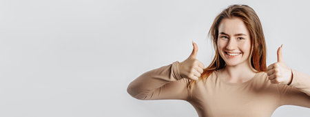 Beautiful young girl smiling and shows thumbs up gesture with two hands on a white isolated background. A woman points to an idea, a place for advertising. Positive brunette in a beige jumper.の写真素材