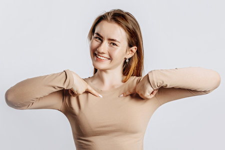 Beautiful young girl smiling and pointing her her fingers at herself on a white isolated background. A woman points to an idea, a place for advertising. Positive brunette in a beige jumper.の写真素材