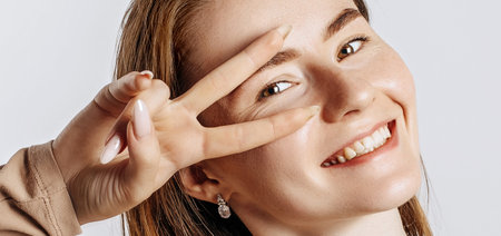 Hey smile you. Young beautiful girl smiles and shows peace victory gesture as fooling around on a white isolated background. A woman points to an idea, a place for advertising. Positive cute brunetteの写真素材