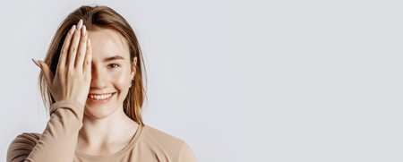 Young beautiful girl smiles and covers half of her face with her hand on a white isolated background. A woman points to an idea, a place for advertising. Positive brunette in a beige jumper.の写真素材