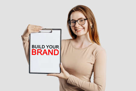 Build Your Brand. Beautiful young business woman wearing glasses holds a clipboard with mock up space isolated on gray backgroundの写真素材