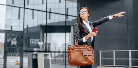 Businesswoman catches a taxi. A woman is waiting for a car and will go to a business meeting. Business concept. Girl with glasses in the city near work.の写真素材
