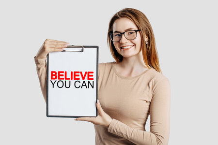 Believe you can. Beautiful young business woman wearing glasses holds a clipboard with mock up space isolated on gray backgroundの写真素材