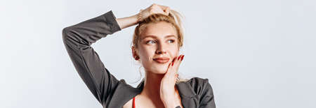 A beautiful blonde girl holds her hair and makes her own hairstyle with the emotion of happiness. Woman dreaming in black jacket and red top on gray isolated background for advertising. Nose piercing.の写真素材