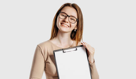 Beautiful young business woman wearing glasses holds a clipboard with mock up blank space isolated on gray background. Achieving career wealth business concept. Online learning coverの写真素材
