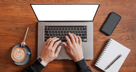 Handsome man working on laptop in the workplace. Businessman typing information on computer at work table with coffee, phone and notepadの写真素材