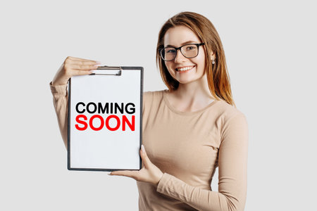 COMING SOON. Beautiful young business woman wearing glasses holds a clipboard with mock up space isolated on gray backgroundの写真素材
