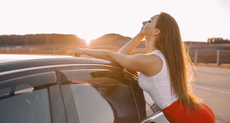 Beautiful young girl near a car in the evening in the sunset sun in an empty parking lotの写真素材