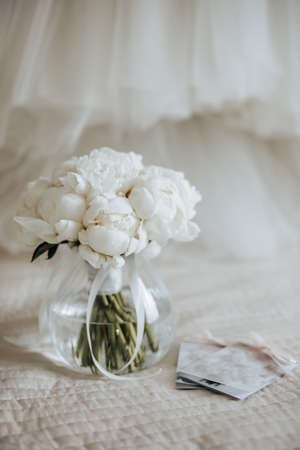 Wedding bouquet of peonies flowers in a vase stands on the bed of the newlyweds with invitationsの写真素材