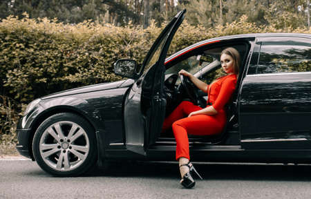 A beautiful young girl in a red overalls sits behind the wheel of a black car on an empty road in the forestの写真素材