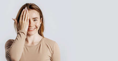 Young beautiful girl smiles and covers half of her face with her hand on a white isolated background. A woman points to an idea, a place for advertising. Positive brunette in a beige jumper.の写真素材
