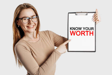 KNOW YOUR WORTH. Beautiful young business woman wearing glasses holds a clipboard with mock up space isolated on gray backgroundの写真素材