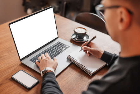 Handsome man working on laptop in the workplace. Businessman typing information on computer at work table with coffee, phone and notepadの写真素材