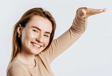 Young beautiful girl smiles and shows big size measure with her hand gesture on a white isolated background. A woman points to an idea, a place for advertising. Positive brunette in a beige jumper.の写真素材