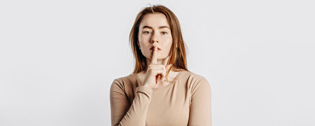 Shh gesture. Young beautiful serious girl holding a finger to her lips on a white isolated background. A woman asks to be silent, a place for advertising. Negative brunette in a beige jumper.の写真素材