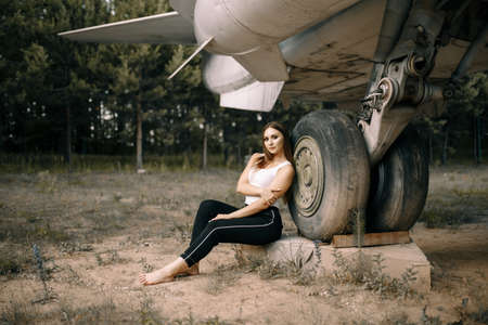 beautiful young brunette girl stands on the background of old military aircraft. Girl in a white shirt and black pants in nature. Military equipment. Portrait half-length. girl posingの写真素材