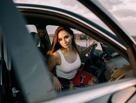 Beautiful young girl driving a car in the evening in the sunset sun on an empty parking lotの写真素材