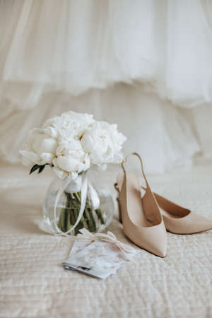 Wedding bouquet of peonies flowers in a vase stands on the bed of the newlyweds with invitations and shoes on the background of the bride dressesの写真素材