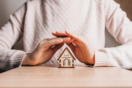 Woman holds and protects a wooden house with her hands with the sun on a light pink background. Sweet homeの写真素材