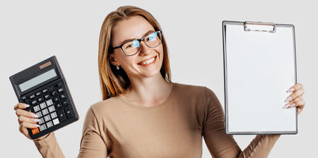 Beautiful young business woman wearing glasses holds calculator and clipboard with mock up blank space isolated on gray background. Achieving career wealth business concept. Cover for advertisingの写真素材