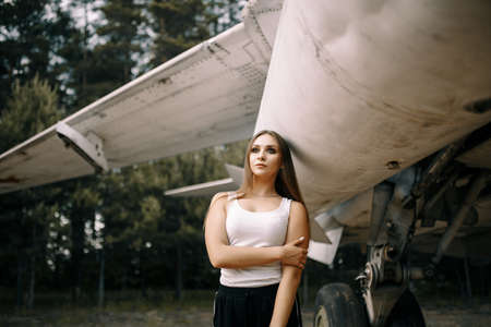 beautiful young brunette girl stands on the background of old military aircraft. Girl in a white shirt and black pants in nature. Military equipment. Portrait half-length. girl posingの写真素材