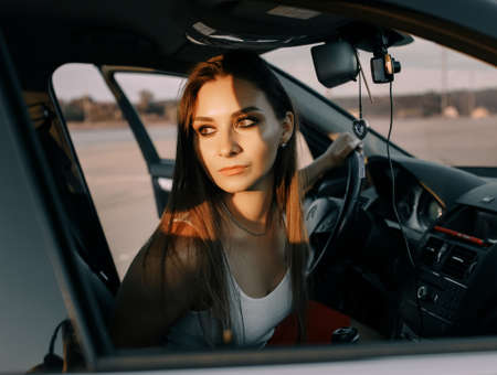 Beautiful young girl driving a car in the evening in the sunset sun on an empty parking lotの写真素材