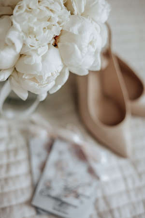 Wedding bouquet of peonies flowers in a vase stands on the bed of the newlyweds with invitations and shoes on the background of the bride dressesの写真素材