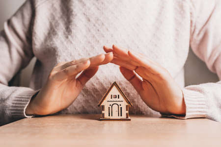Woman holds and protects a wooden house with her hands with the sun on a light pink background. Sweet homeの写真素材