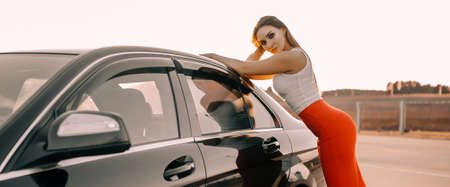Beautiful young girl near a car in the evening in the sunset sun in an empty parking lotの写真素材