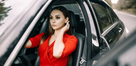 A beautiful young girl in a red overalls sits behind the wheel of a black car on an empty road in the forestの写真素材