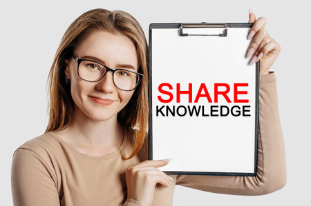 SHARE KNOWLEDGE. Beautiful young business woman wearing glasses holds a clipboard with mock up space isolated on gray backgroundの写真素材