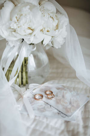 Wedding bouquet of peonies flowers in a vase stands on the bed of the newlyweds with invitations and rings on the background of of a veil of the brideの写真素材