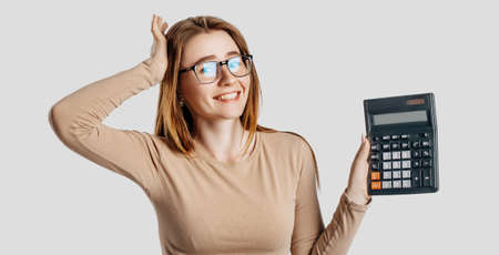 Beautiful young business woman wearing glasses holds calculator isolated on gray background. Achieving career wealth business accounting concept. Cover for advertisingの写真素材
