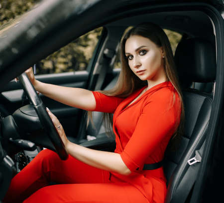 A beautiful young girl in a red overalls sits behind the wheel of a black car on an empty road in the forestの写真素材